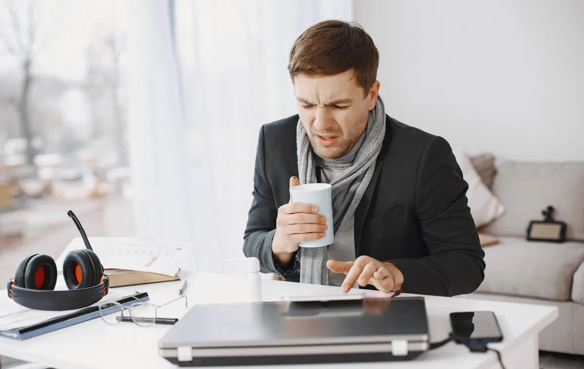 Un salarié en manteau et écharpe réchauffe ses mains autour d'une tasse devant son ordinateur, dans un bureau où il fait trop froid