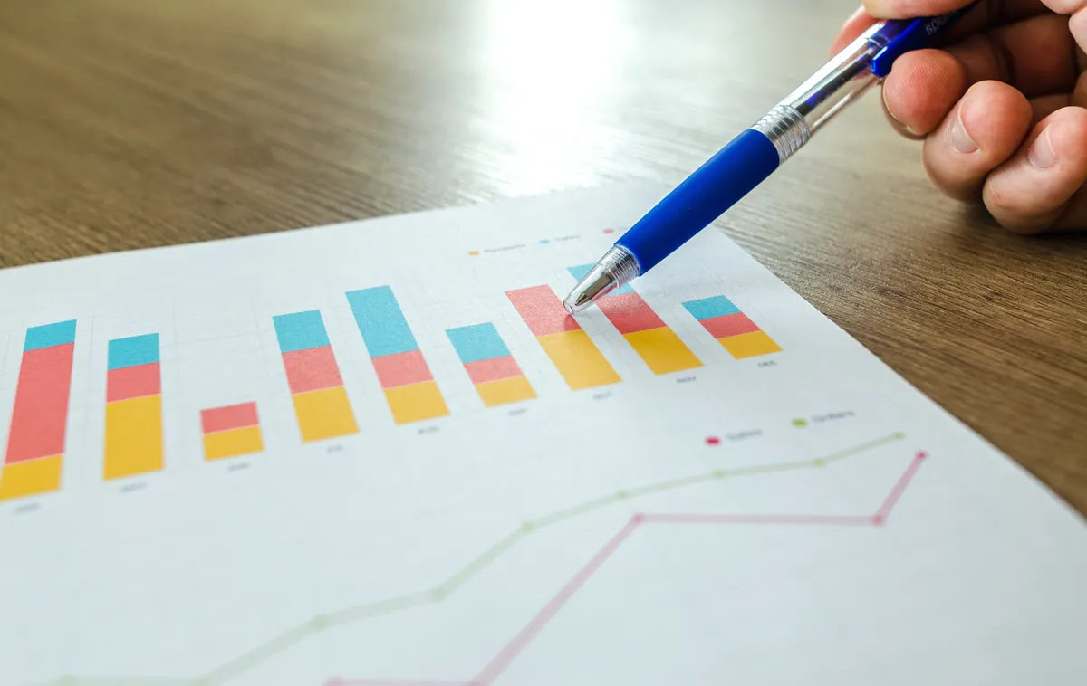 Une main tient un stylo bleu au-dessus d'un graphique en barres et en courbes imprimé, posé sur un bureau en bois