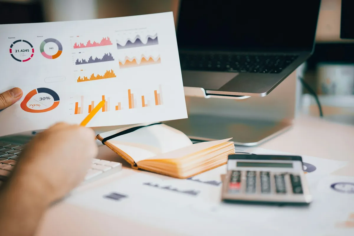 Un rapport imprimé couvert de graphiques et de diagrammes circulaires, tenu au-dessus d'un bureau où se trouvent un ordinateur portable et une calculatrice
