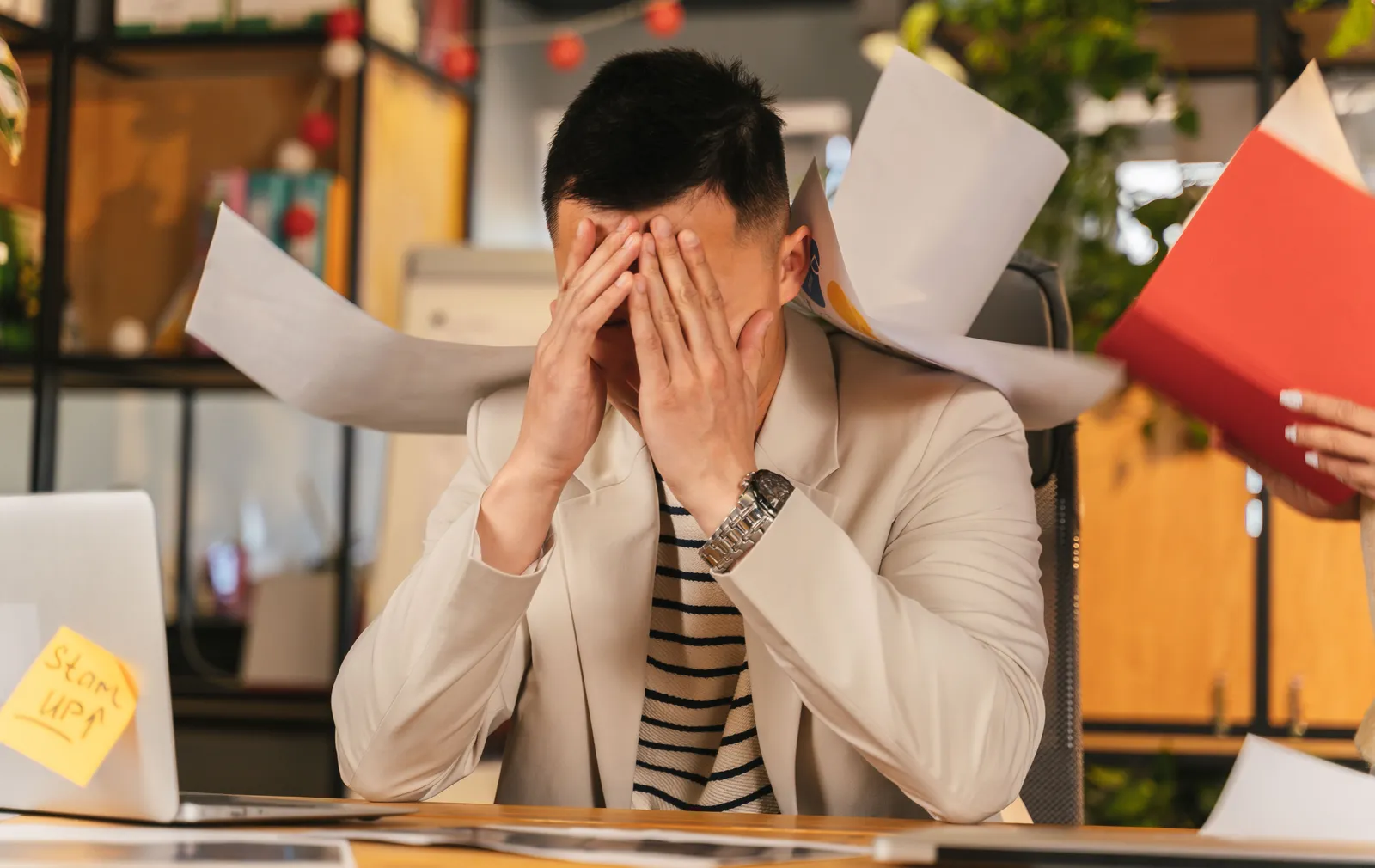 Un collaborateur assis à son bureau se prend la tête dans les mains, submergé par les documents qui volent autour de lui