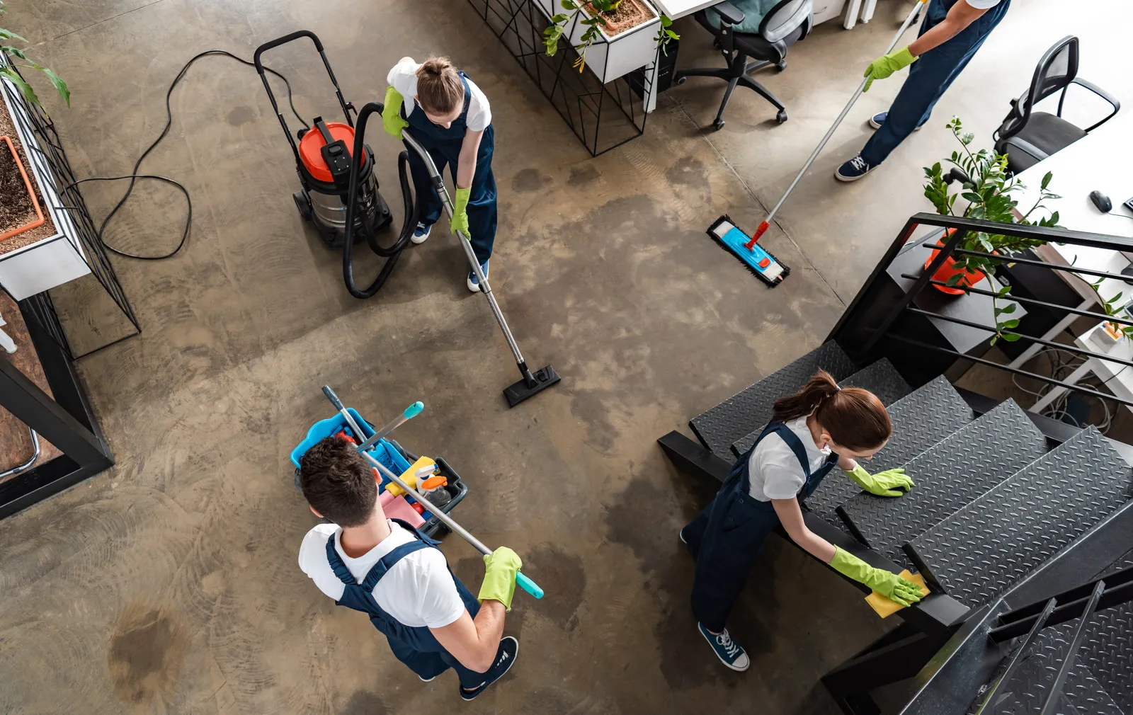 Trois agents de propreté en salopette nettoient un open space, vus en plongée : passage de l'aspirateur, du balai et nettoyage d'une table